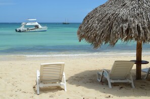 A beach with two white lounge chairs, a thatched umbrella, and a boat in the water.