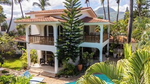 Un bâtiment à deux étages doté d’un balcon, entouré d’une végétation tropicale et d’une piscine.