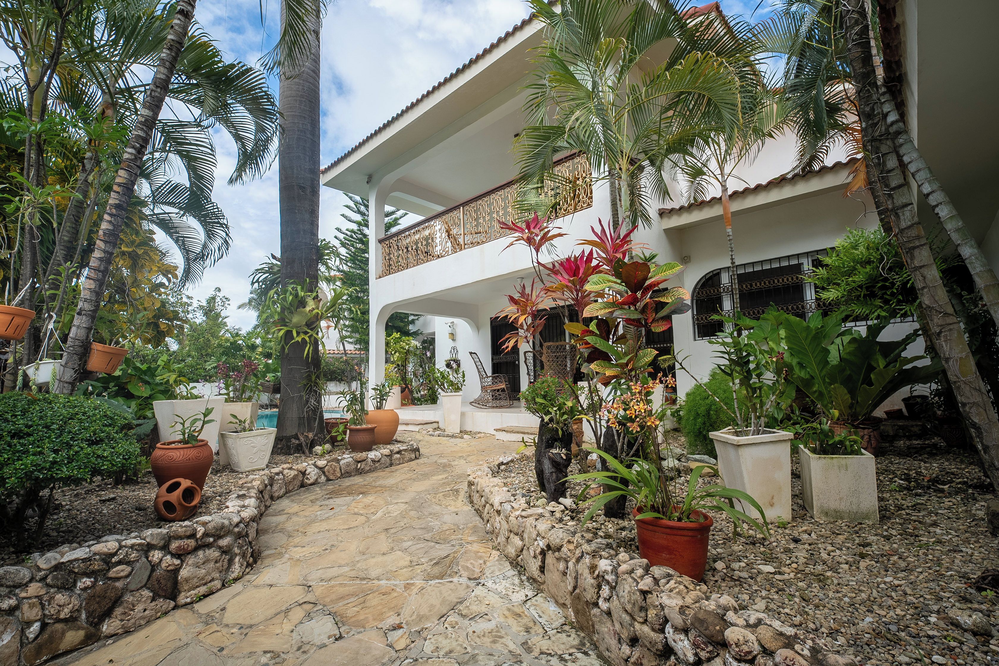 A courtyard with a stone pathway, surrounded by potted plants and palm trees, leading to a white building with a balcony.
