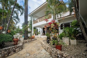 Une cour avec un chemin en pierre, bordée de plantes en pot et de palmiers, qui mène à un bâtiment blanc doté d’un balcon.