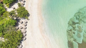 Vue aérienne d’une plage aux eaux turquoise et limpides, au sable blanc et à la végétation luxuriante.