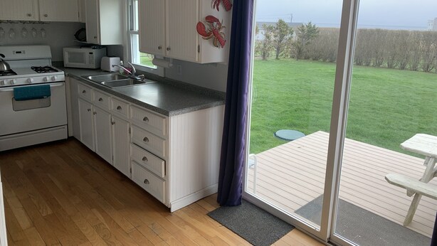A kitchen with white cabinets, a stove, and a microwave. There is a large sliding glass door leading to a deck with a picnic table. - Ocean View Home With Large Yard, A/c, Hot Tub, Walk To Sandy Beach, Pets Ok (Narragansett)