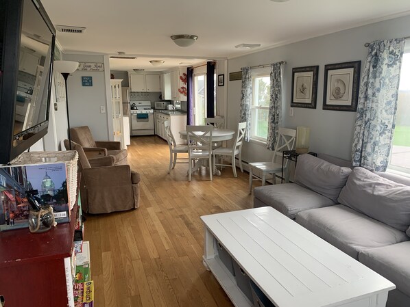 A spacious living room with a white sofa, a dining table with chairs, and a kitchen in the background. - Ocean View Home With Large Yard, A/c, Hot Tub, Walk To Sandy Beach, Pets Ok (Narragansett)