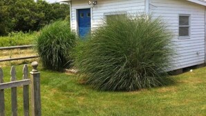 A small, single-story white building with a blue door, surrounded by grass and bushes. - Ocean View Home With Large Yard, A/c, Hot Tub, Walk To Sandy Beach, Pets Ok (Narragansett)