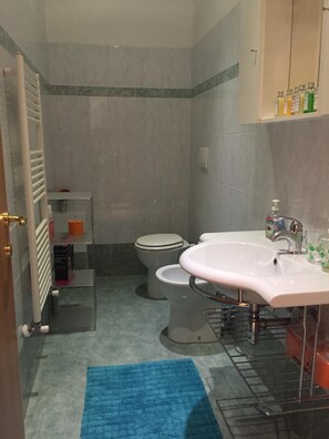 Un baño con dos inodoros, un lavamanos, cabina de ducha de vidrio, alfombra azul y un estante con artículos de tocador.