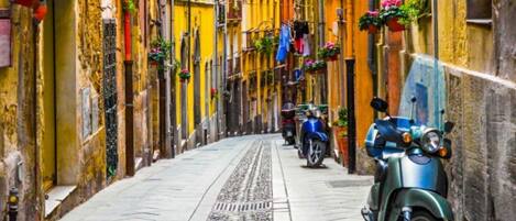 Una calle estrecha flanqueada por edificios, algunos con balcones y plantas colgantes. Hay scooters estacionados y una bicicleta.