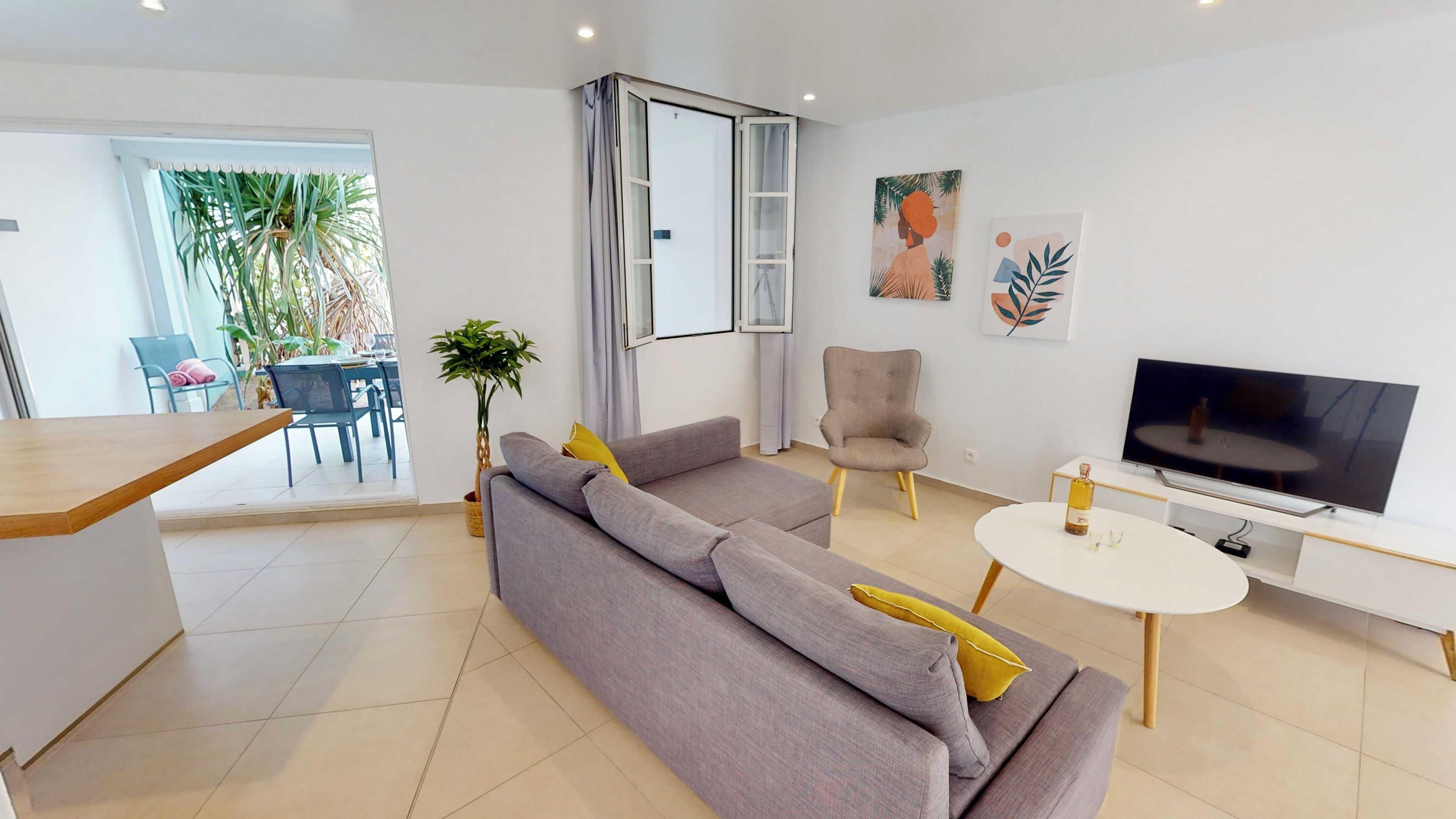 Una sala de estar moderna con un sofá gris, una mesita redonda, un televisor y un comedor con sillas y una mesa a la vista a través de un umbral abierto.