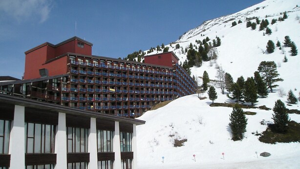 A multi-story hotel with a red and blue facade, located at the base of a snow-covered mountain. - Arc 2000, studio direct access to  tracks, South balcony (Bourg-Saint-Maurice)