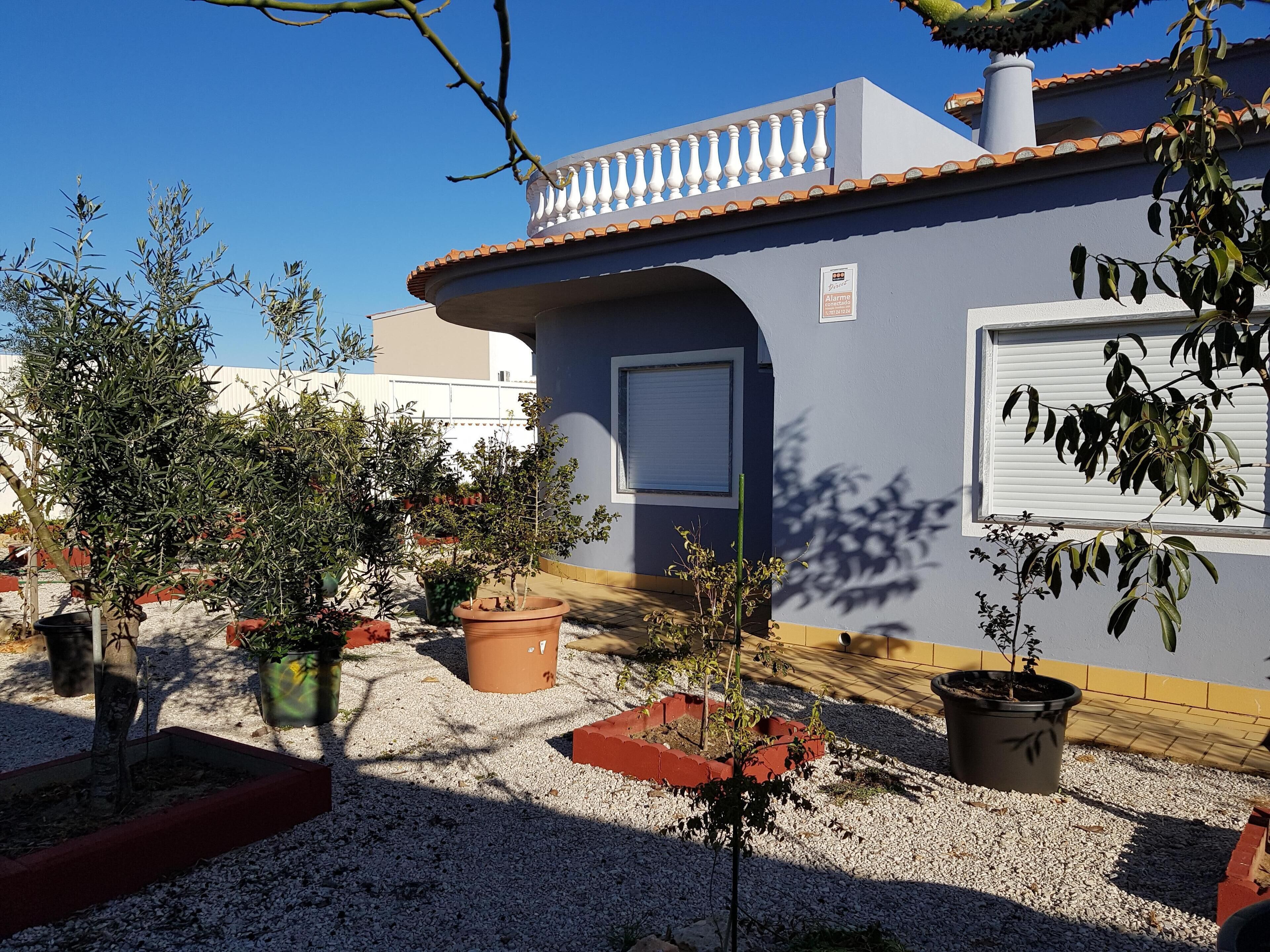 Um edifício com fachada cinzenta, telhado de telhas e varanda com balaústres brancos. Há plantas em vasos na frente do edifício.