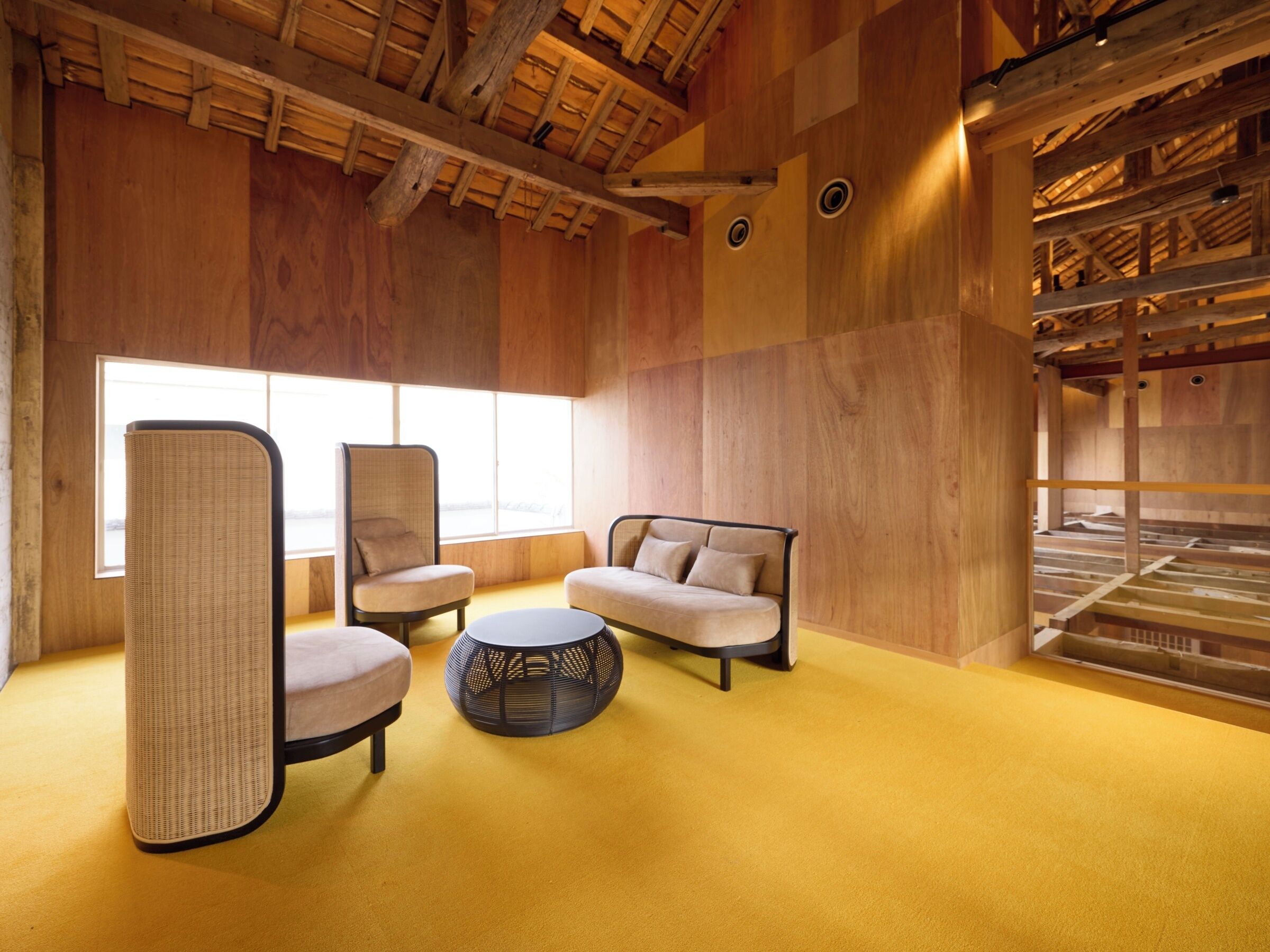 Una habitación con techo alto, vigas de madera y un ventanal grande. Hay dos sillones y un sofá con cojines. Una mesa redonda está en el centro.