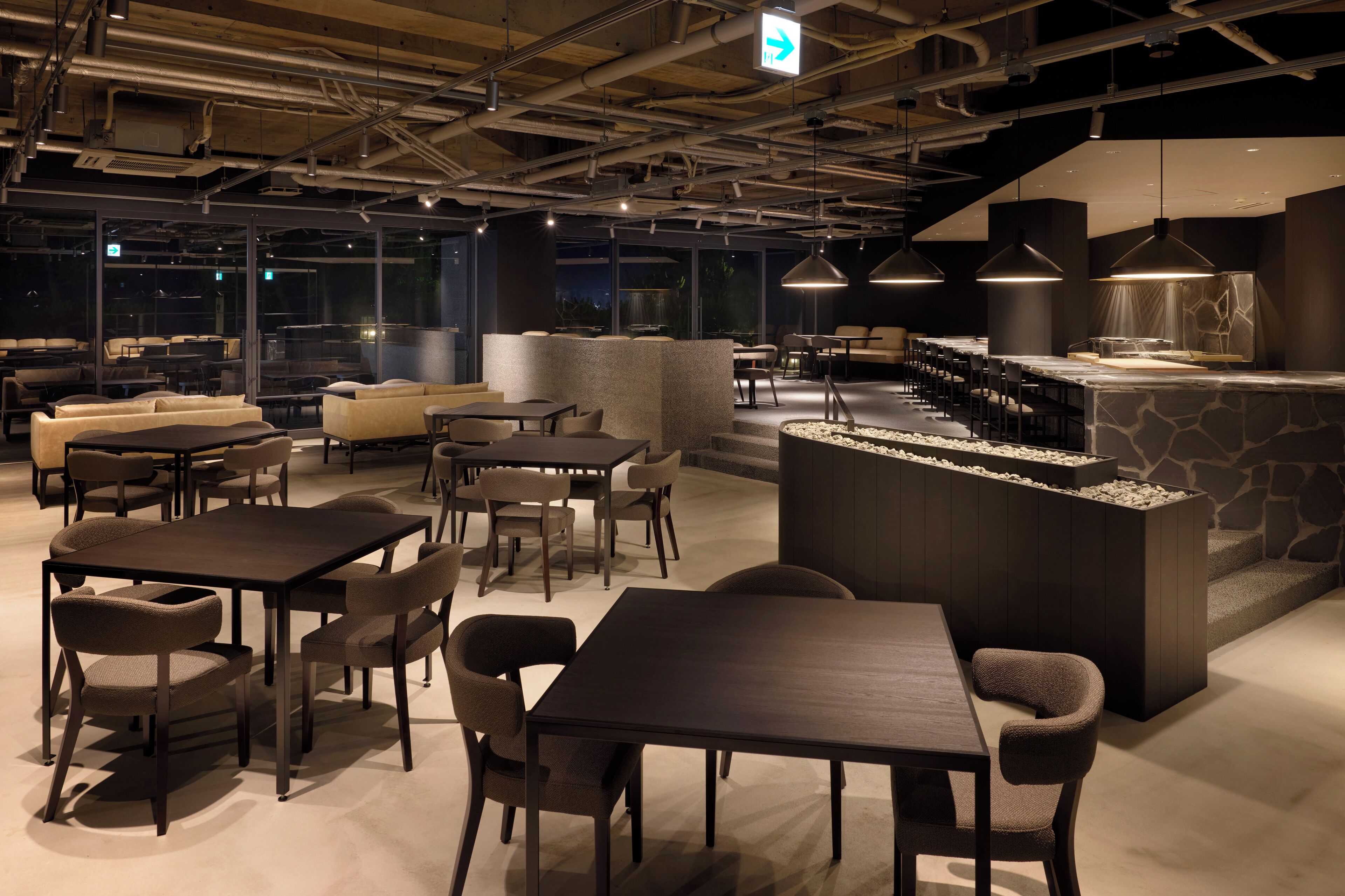 Uma área de estar moderna com diversas opções de assento, incluindo mesas e sofás, um bar com bancada de pedra e vista para o exterior através de amplas janelas.