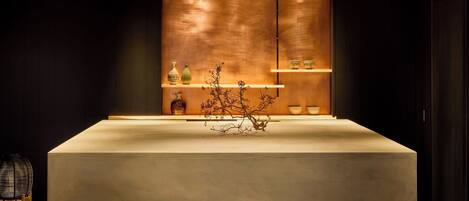 Un área de recepción moderna y minimalista, con un mostrador central, estanterías con objetos decorativos y una pared de tonos oscuros.
