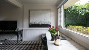 Una habitación con un cuadro abstracto grande en la pared, un televisor sobre una mesa negra, un florero con flores y una ventana con vistas a la vegetación.