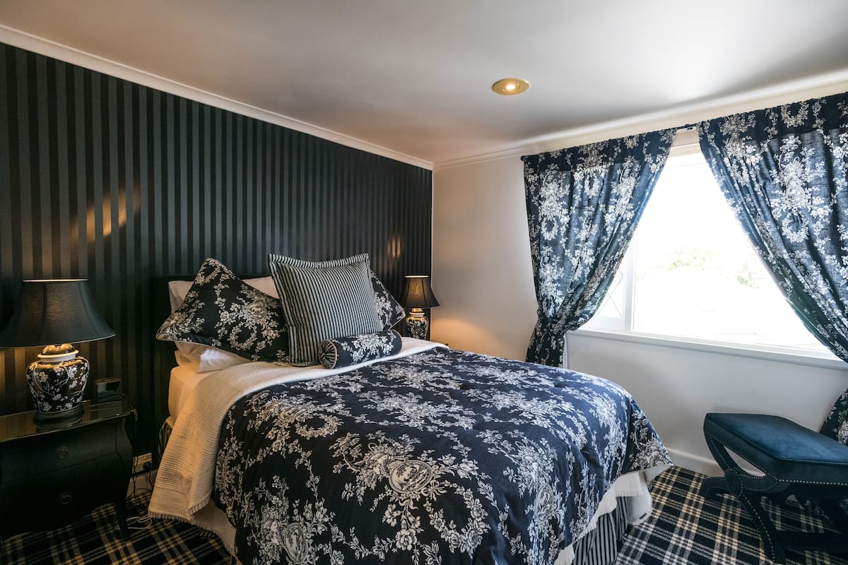 Une chambre à coucher avec un lit, une table de chevet, une lampe, un banc et une fenêtre avec des rideaux.