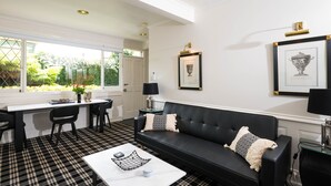 Una sala de estar moderna con un sofá de cuero negro, una mesa de centro blanca y un suelo a cuadros blanco y negro. Hay una zona de comedor con una mesa blanca y sillas negras, y una ventana con vista a la vegetación.