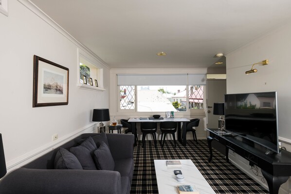 A modern living room with a sofa, coffee table, and TV. There is a dining area with chairs and a table. The room has a checkered floor and large windows. - Battery Point Boutique Accommodation (Battery Point)