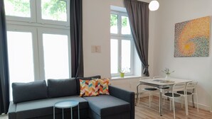 Una sala de estar moderna con un sofá seccional gris, una mesita redonda, una mesa de comedor blanca con sillas y un ventanal con cortinas.