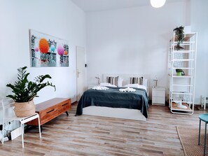 A modern bedroom with a wooden floor, a bed with a dark green bedspread, white pillows, a white nightstand, a white shelf unit, and a potted plant.