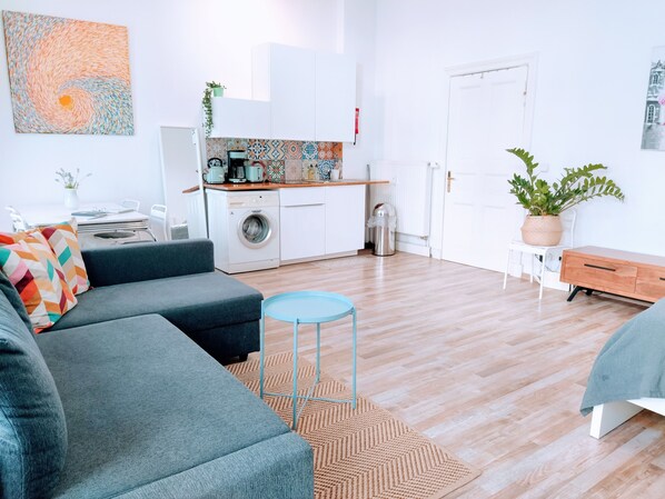 A modern living room with a grey sofa, a small round table, and a kitchen area with a washing machine and a coffee maker.