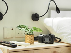 Een netjes opgemaakt bed met witte lakens, een nachtkastje met een camera, een afstandsbediening en een potplant.