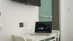 A small, white room with a single white chair and table, a laptop, a flat-screen TV, and a green wall. - Ukkei Inn (Kowloon)