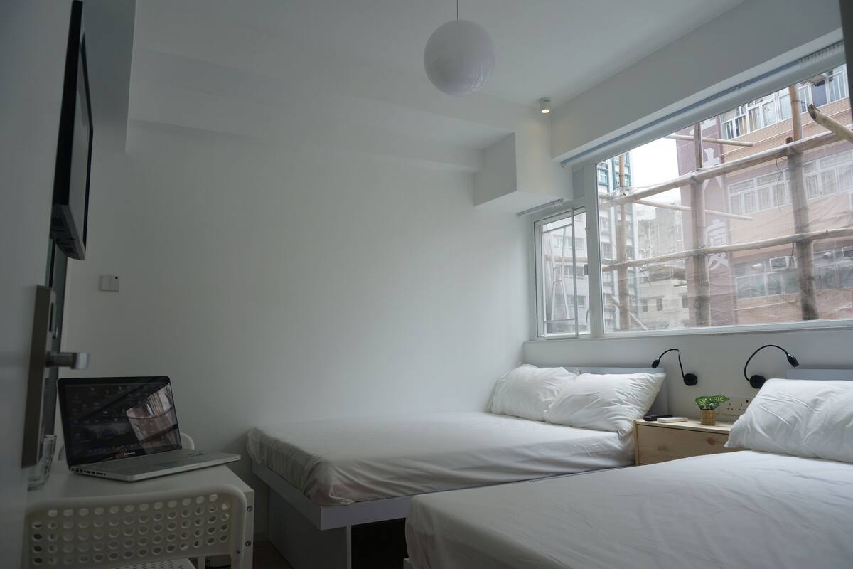 A room with two beds, a laptop on a small table, and a window with a view of buildings.