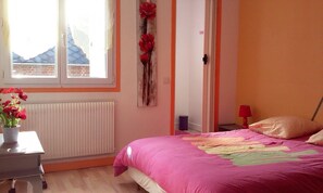 A bedroom with a pink bedspread, a radiator, a window with a view of buildings, and a door with a red flower decoration.