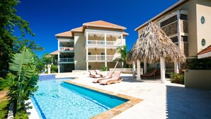A pool area with lounge chairs, a thatched-roof structure, and a multi-story building with balconies. - Lawson Rock - Angelfish 100 (Roatan)