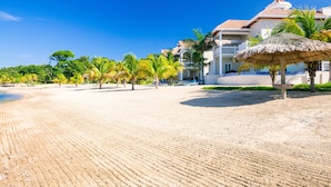 A beach with a thatched umbrella, palm trees, and a building with balconies. - Lawson Rock - Angelfish 100 (Roatan)