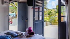 Ein Schlafzimmer mit einem Bett, einem Handtuch mit Blumenmotiv und Blick auf einen Balkon mit Pflanzen sowie ein Gebäude im Hintergrund.
