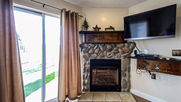 A living room with a stone fireplace, a flat-screen TV mounted above it, and a sliding glass door leading to a balcony. - Inn Building Condo#113 - Ground Level, Pool Side (Garden City)