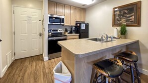 A modern kitchen with wooden cabinets, stainless steel appliances, a granite countertop, and a bar area with stools. - Inn Building Condo#113 - Ground Level, Pool Side (Garden City)