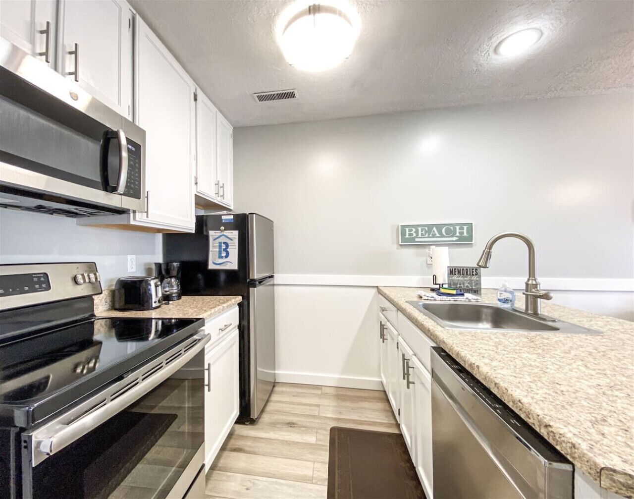 A modern kitchen with stainless steel appliances, white cabinets, a granite countertop, and a 'Beach' sign on the wall.