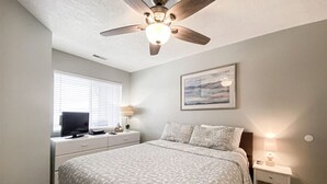 A bedroom with a bed, a ceiling fan, a TV, and a bedside table with a lamp. - Inn Building Condo #203- Remolded with Lake View! (Garden City)