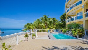 A pool area with a view of the ocean, surrounded by palm trees and a building with balconies. - Villa Del Playa #2 (Roatan)