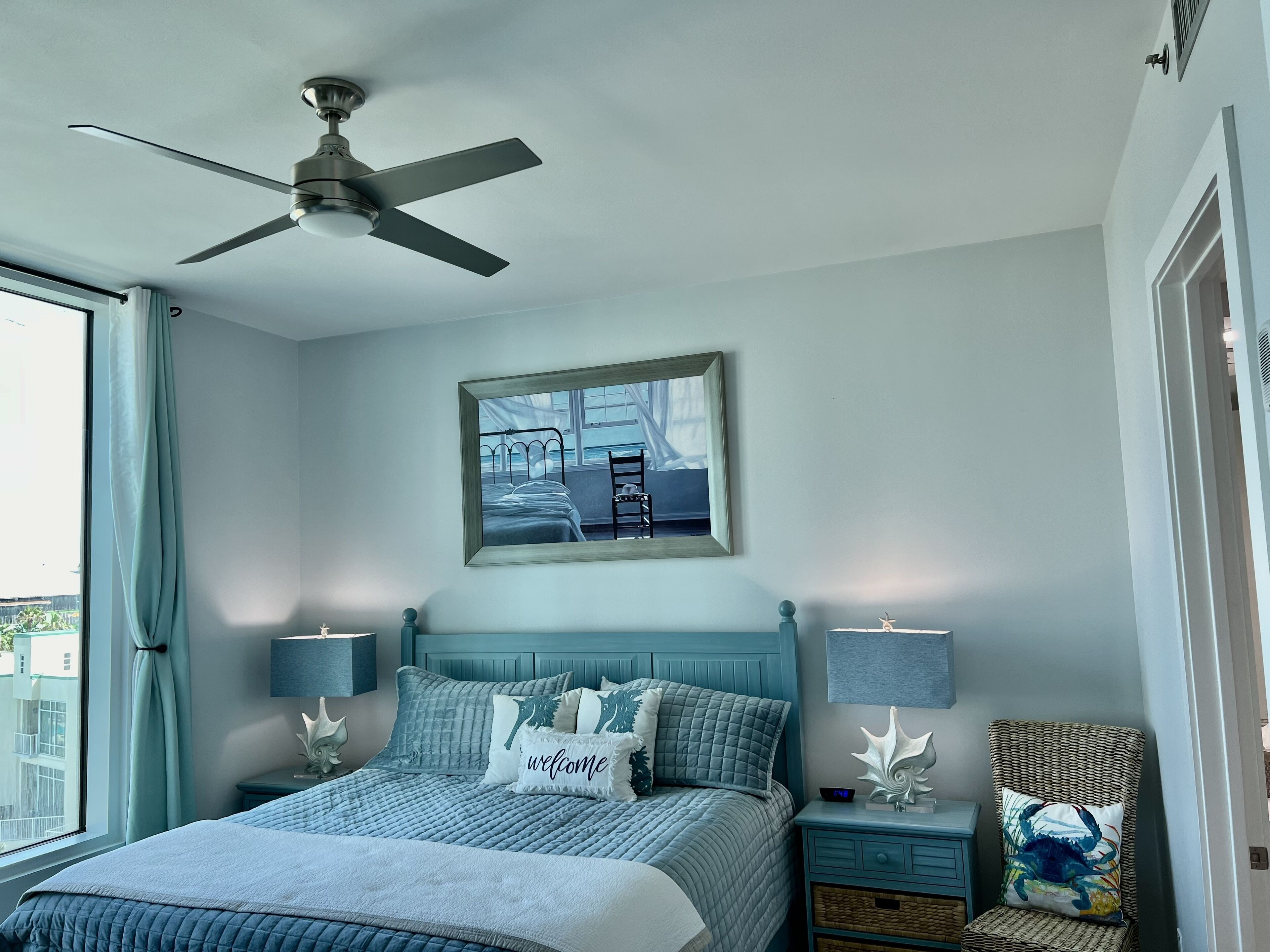 A bedroom with a bed, a ceiling fan, a window with curtains, a nightstand with a lamp, and a wall-mounted picture.