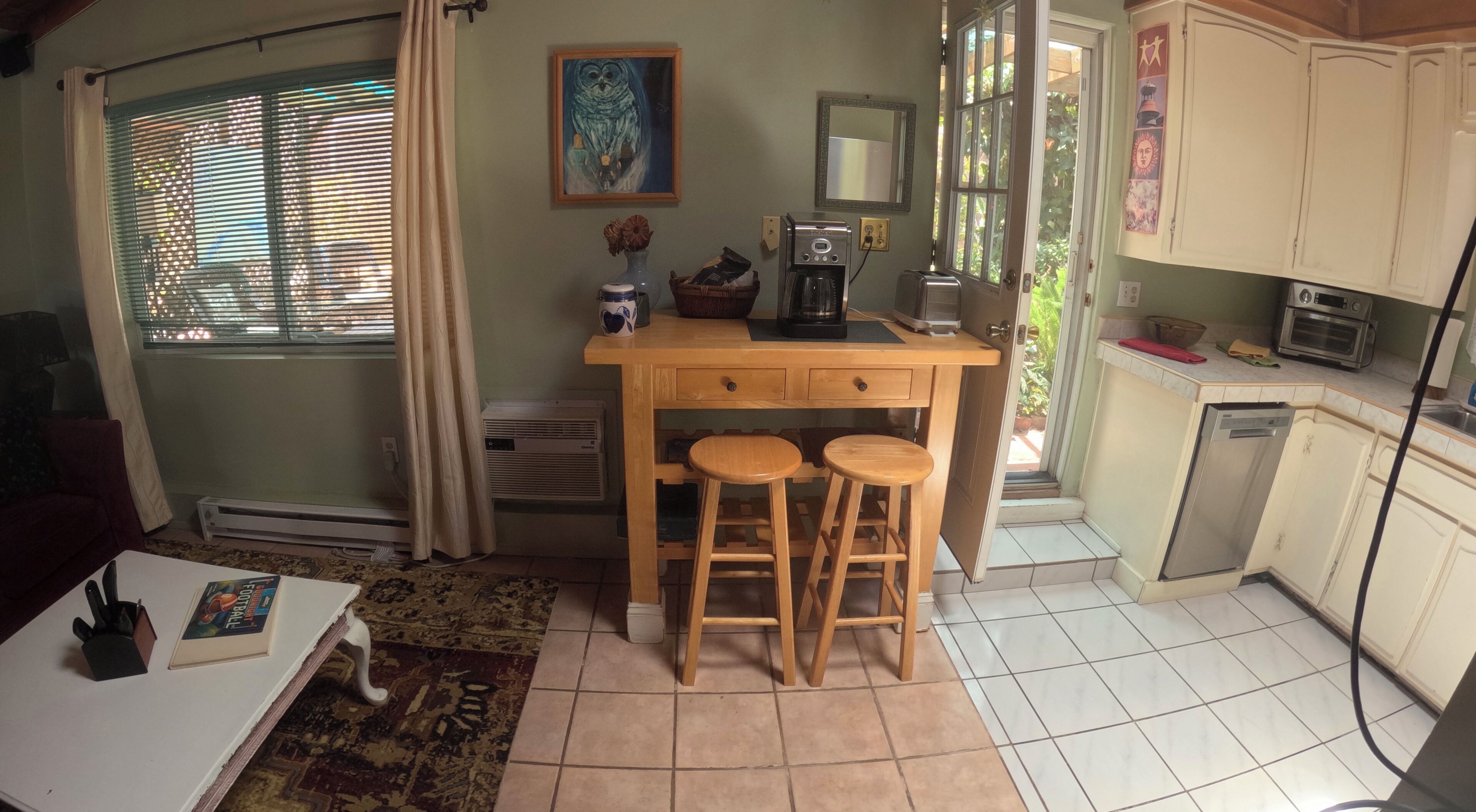 Eine kompakte Küche mit Kaffeemaschine, Toaster und kleinem Kühlschrank. Am Tresen stehen zwei Hocker, ein Fenster mit Jalousien und ein Bild an der Wand.