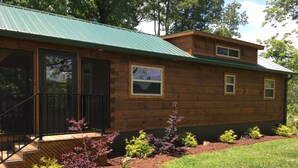 A wooden cabin with a green roof, a front porch, and a small garden with various plants.