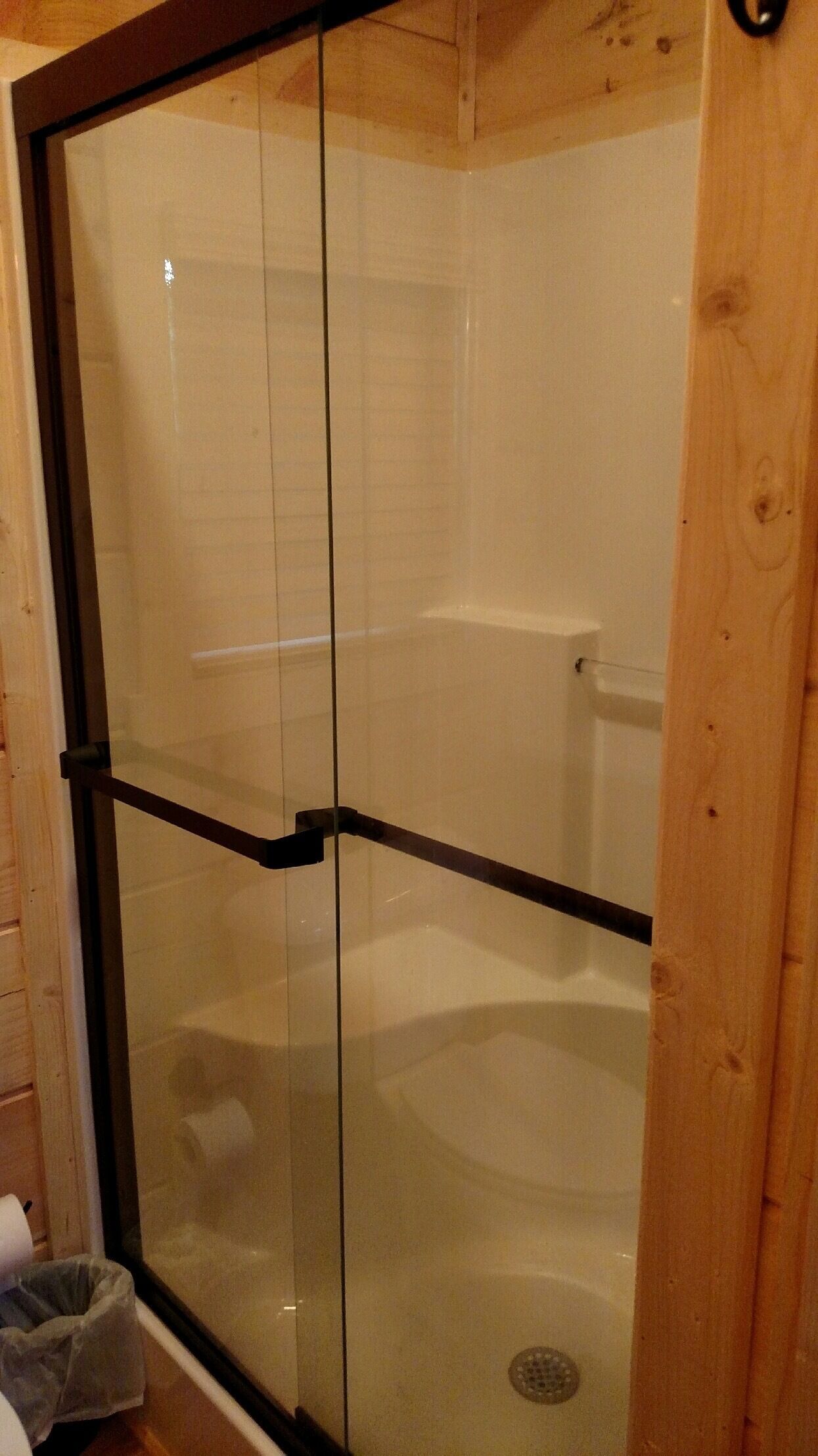 Ducha con cerramiento de vidrio, estructura de madera, pared con azulejos blancos y desagüe visible.