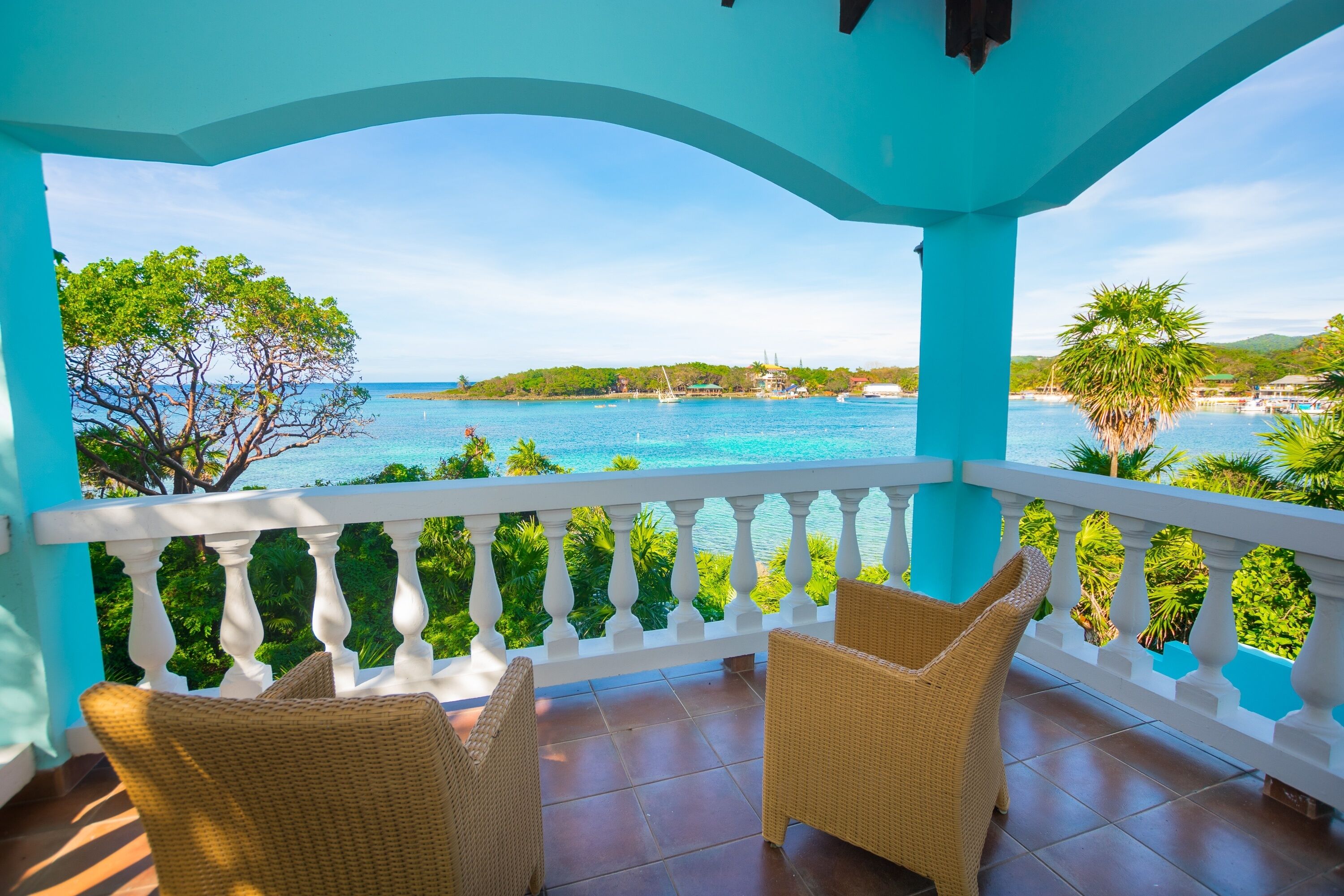 A balcony with wicker chairs overlooking a turquoise sea and a coastal town.
