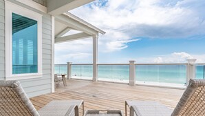 A covered deck with wicker lounge chairs and a table, overlooking a beach with clear blue water.