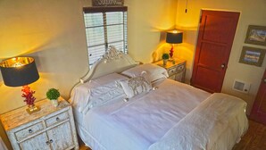 Un dormitorio con una cama, dos mesitas de noche con lámparas, una mesita de madera, una puerta y una ventana con persianas.