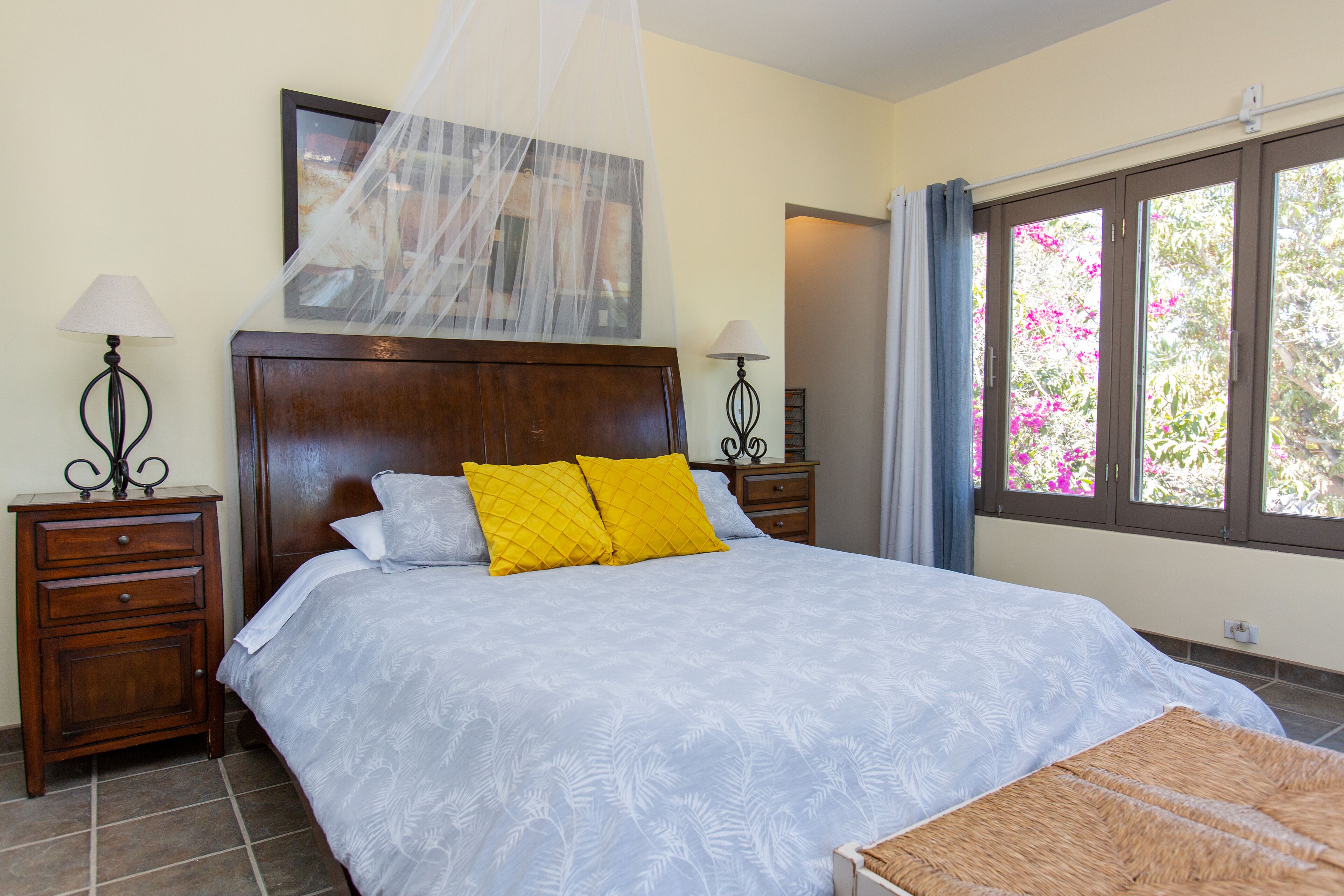 A bedroom with a large bed, two nightstands, a wooden headboard, a canopy, and a window with a view of pink flowers.