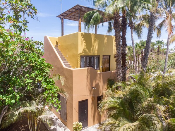 Un edificio de dos plantas con una escalera que conduce al nivel superior, rodeado de exuberante vegetación y palmeras.