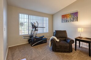 A living room with a treadmill, a leather armchair, a small table with a lamp, and a colorful painting on the wall.