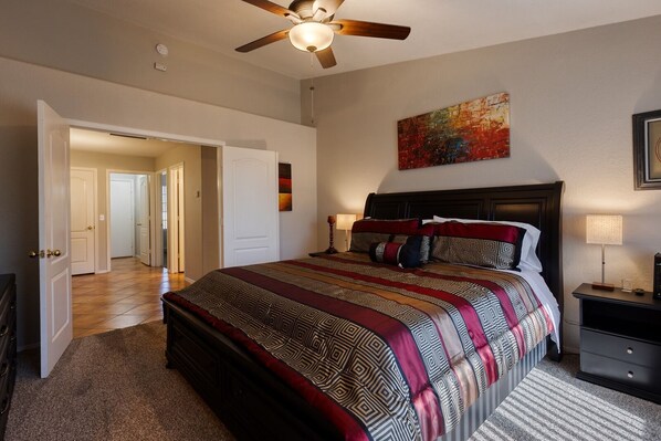 A bedroom with a large bed, a ceiling fan, a nightstand with a lamp, and a door leading to another room.