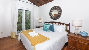 A hotel room with a bed, a wooden headboard, a nightstand with a lamp, a mirror, and a view of greenery through a window.