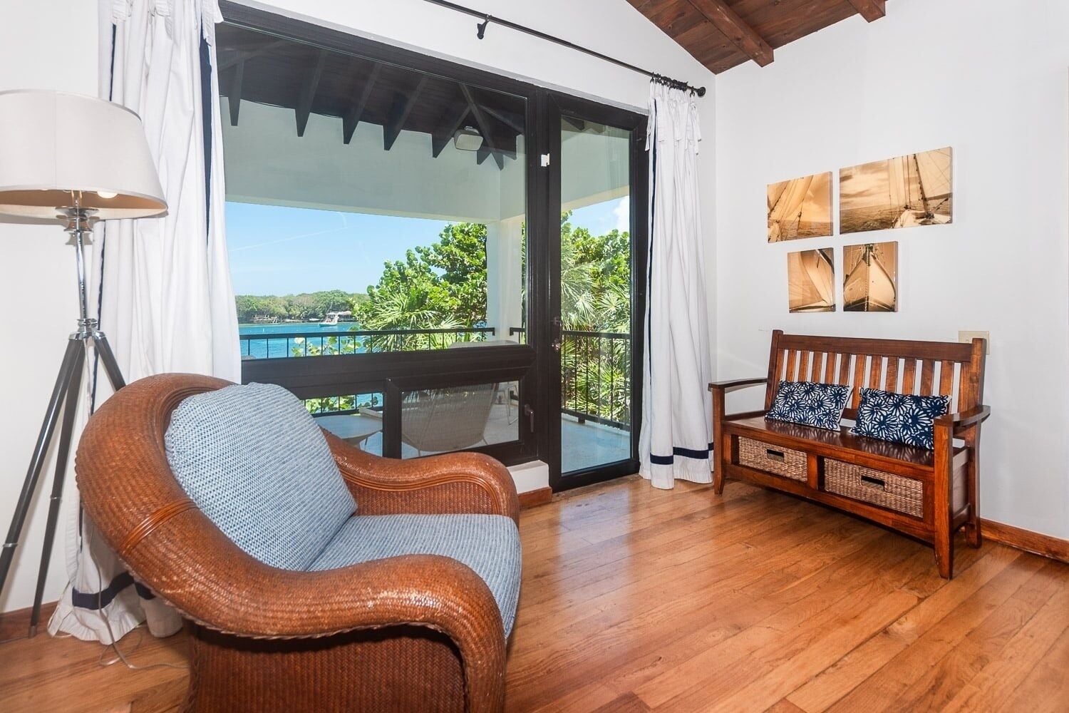 A room with a wicker chair, a wooden bench with patterned cushions, a floor lamp, and a view of a body of water through a glass door.