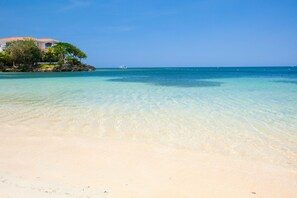 Une plage à l’eau turquoise et limpide, un bâtiment blanc doté d’un balcon, et un bateau aperçu au loin.