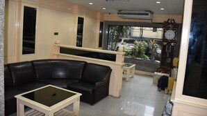 A lobby with a black leather sofa, a glass-top coffee table, and a wooden bench. There is a large clock on the wall and a potted plant in the background. - Woodlands Inn (Bangkok)